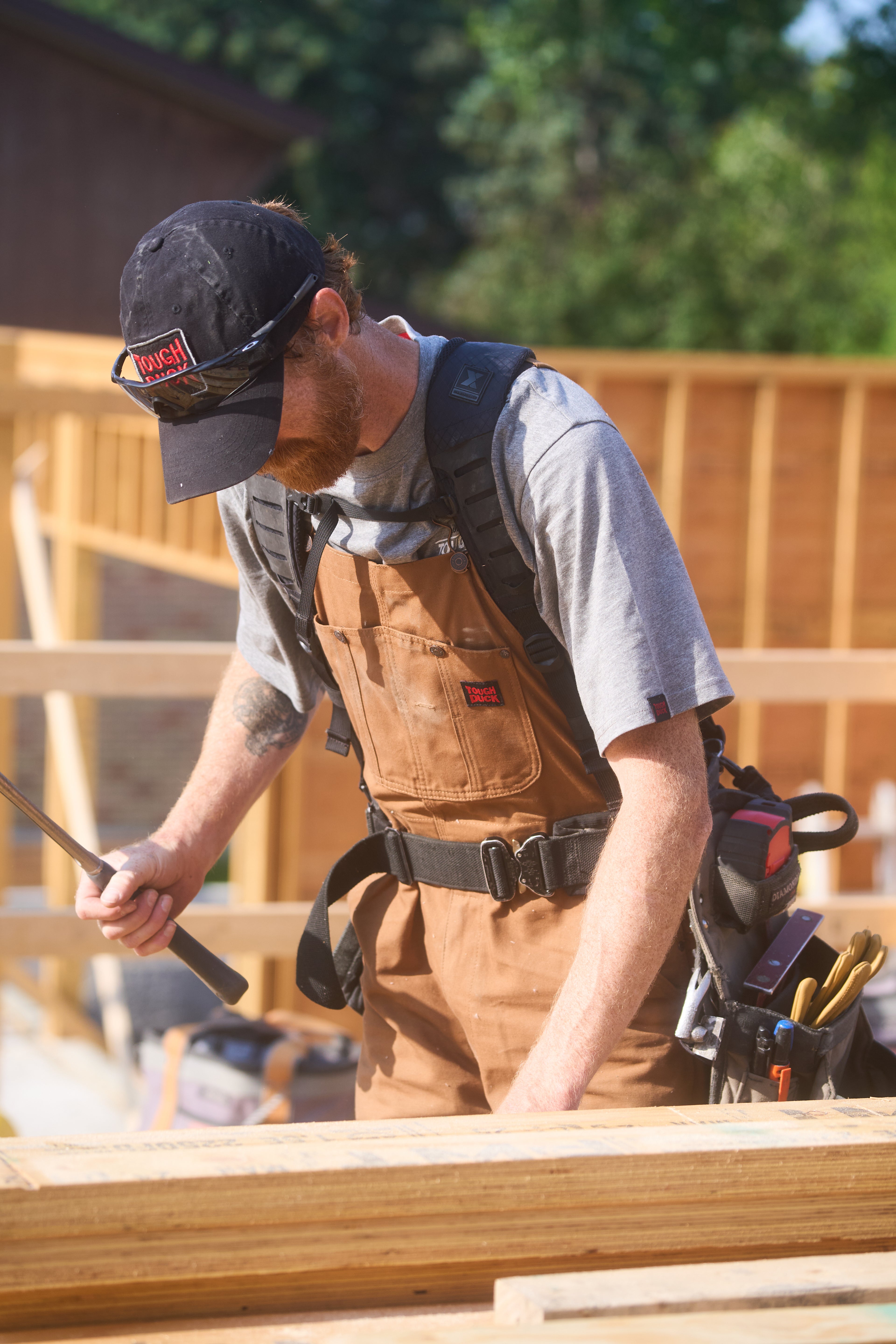 Carpenter wearing Tough Duck brown bib overalls and ball cap framing on construction jobsite
