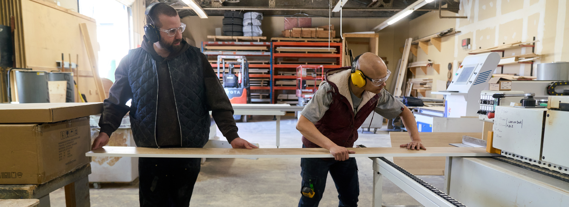 Two men in a workshop, one holding a long piece of wood.