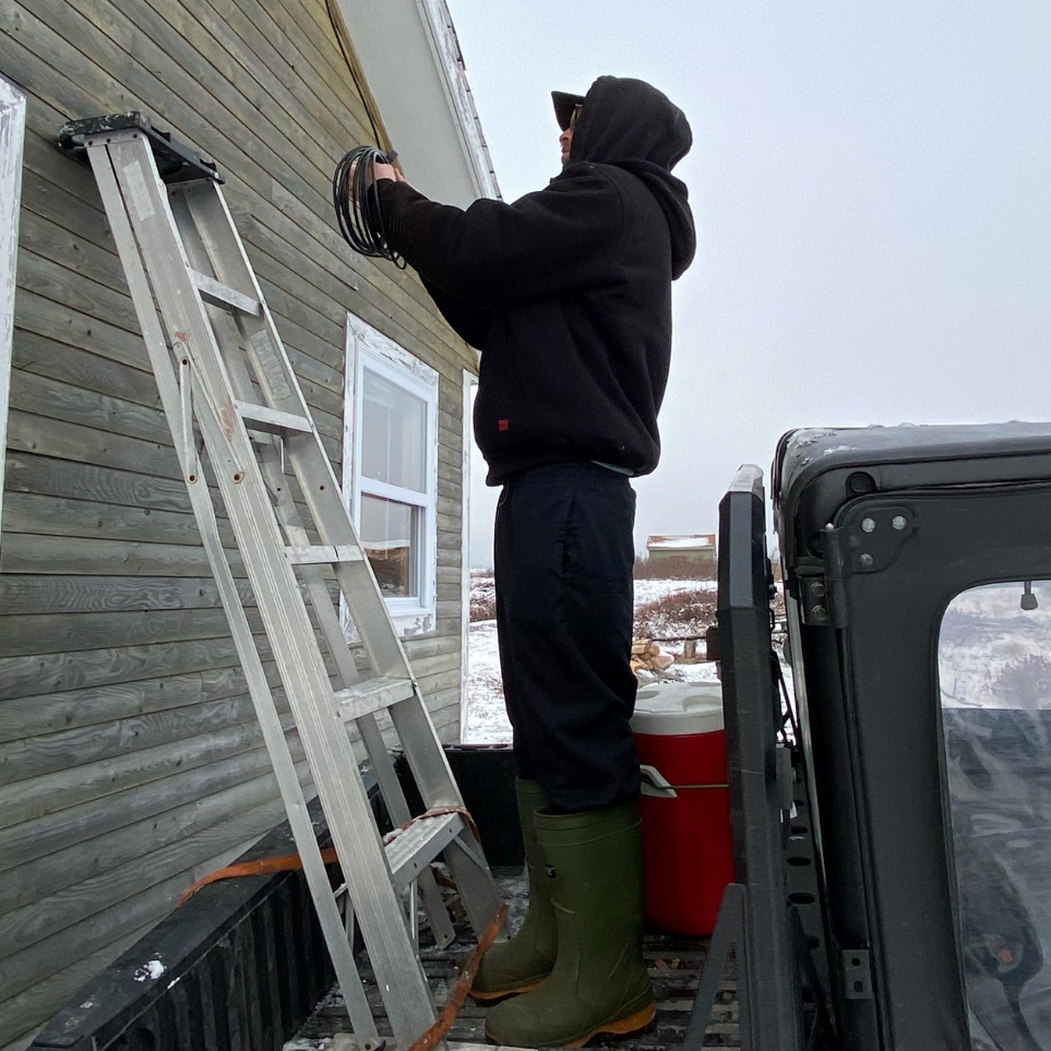 Person on a ladder working on a roof with a vehicle and cooler nearby, wearing a black hoodie WJ08